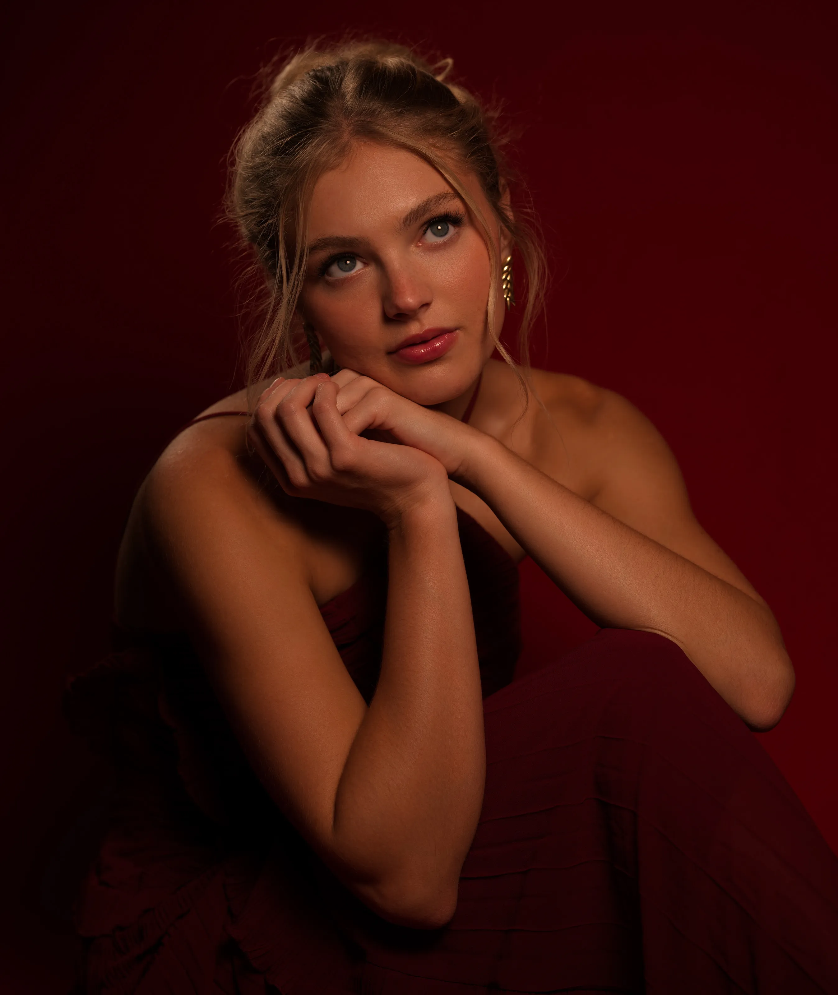 Blonde woman in a deep red dress, seated on a dark red backdrop.