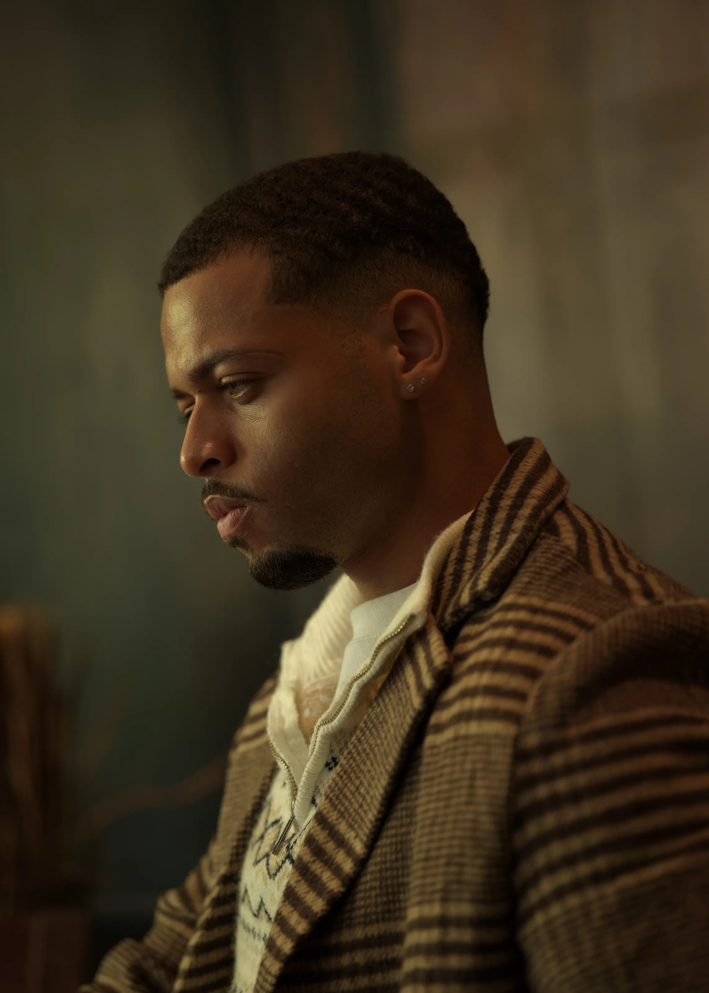 Man in profile in a brown striped coat on a muted green backdrop.