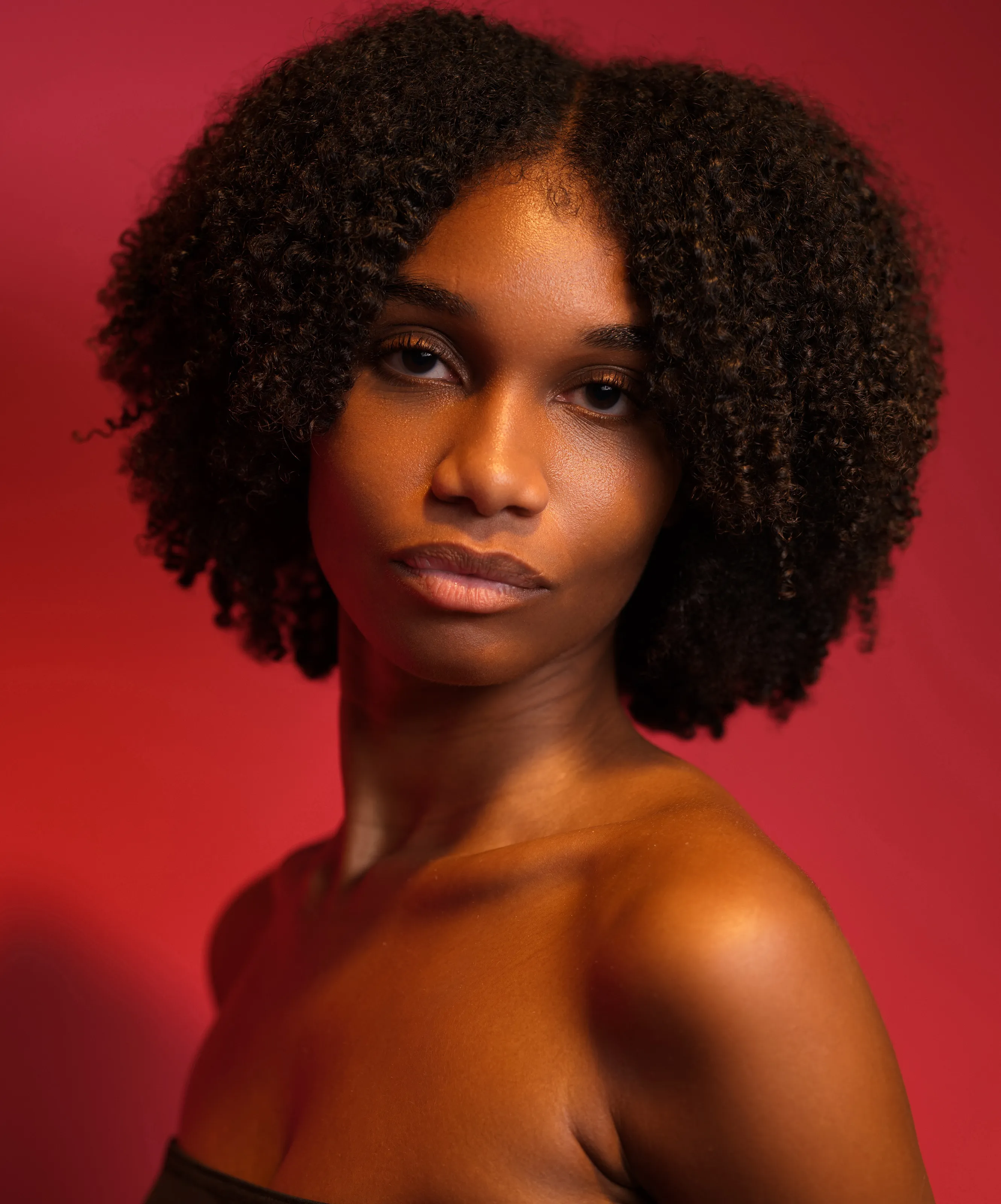 Woman with curly hair and bare shoulders on a vivid red backdrop.